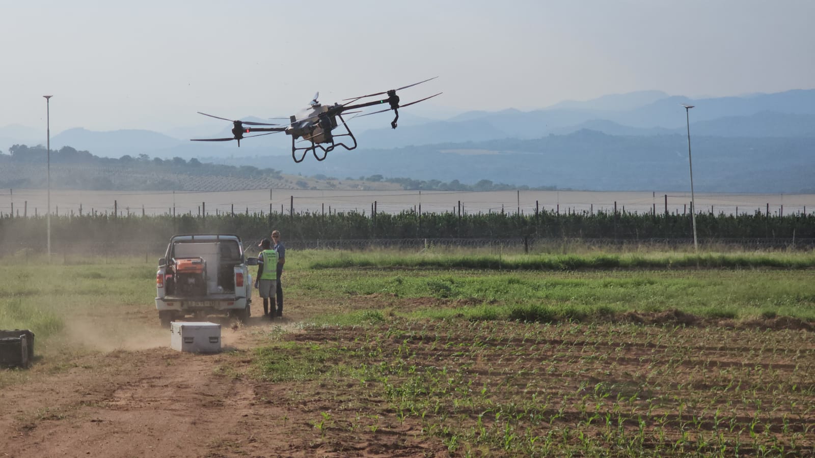 Spray drone in flight above a field crop with a support vehicle and operators on the dirt track